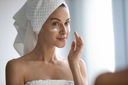 Close up head shot beautiful woman with healthy perfect smooth skin applying cream on cheeks, pretty young female with white bath towel on head looking in mirror in bathroom, skincare and spaの写真素材