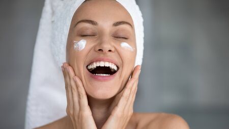 Close up excited beautiful woman with toothy healthy smile applying moisturizing cream on face,...