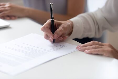 Close up view female hand holding pen signing legal paper seated at desk indoors, client receives...