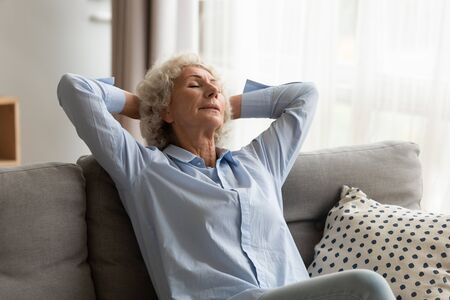 Older woman relaxing at home in modern living room, sleeping on comfortable couch with hands behind...