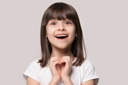 Close up head shot portrait cheerful excited small 6 years old girl looking at camera, showing heart...
