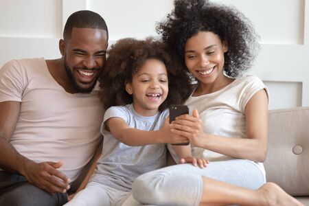 Preschool small daughter having fun use smartphone showing new game funny video to parents, full african family enjoy free time together make selfie photo, modern technology and entertainment conceptの写真素材