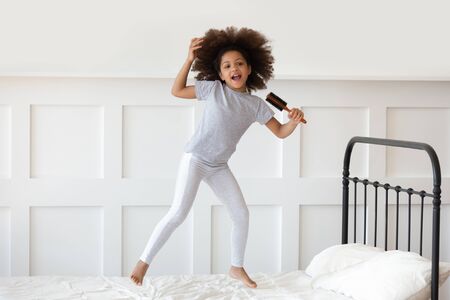 Full-length of little african girl in night-wear awoken jumping on bed holding hair brush like microphone open mouth singing imitates imagines herself a real singer, hobby mood pastime at home conceptの写真素材