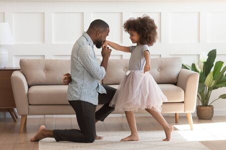 In cozy living room african dad got down on one knee kisses hand of daughter princess wearing fluffy pink skirt, chivalrous gesture, courtesy and politeness, devotion admiration, good manners conceptの写真素材