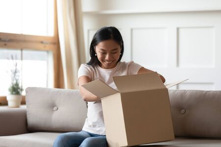 Overjoyed young Asian woman sit on couch open cardboard box with Internet order, excited millennial...