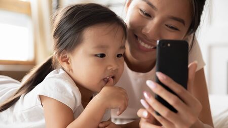 Smiling Asian millennial mom or nanny relax with little biracial girl child using cellphone...