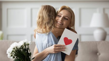 Head shot happy mommy embracing little preschool child girl, thanking for congratulations, handmade card with love symbol and flowers. Smiling blonde mother excited by daughters present at home.の写真素材