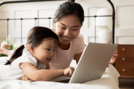 Happy young Asian mom and little ethnic daughter lying in bed at home watch video on modern laptop, smiling Vietnamese mother or nanny relax in bedroom with small biracial girl child using computerの写真素材
