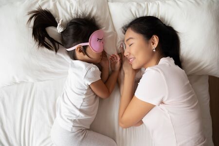 Top view of happy young Asian mom and little daughter lying in comfortable bed sleep daydream at home together, Vietnamese mother and small biracial girl child asleep take nap dream in cozy bedroomの写真素材