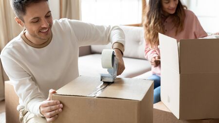 Smiling Caucasian husband packing together with wife sealing carton boxes using adhesive tape, young...