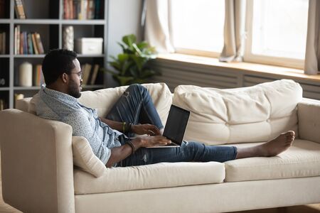 Young african man using laptop studying or working in app typing on computer lounge on sofa, millennial adult afro american male user holding notebook computer relax on couch in living room at homeの写真素材