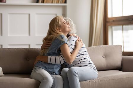 Loving two generations family embracing on couch, reconciling after quarrel at home. Peaceful young grown up daughter cuddling middle aged hoary mother, enjoying tender sweet moment together.の写真素材