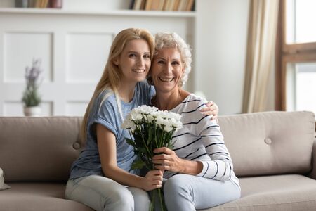 Portrait of young smiling blonde grown up daughter embracing middle aged retired mother, holding flowers, sitting together at home. Happy two generations family celebrating birthday, posing for photo.の写真素材