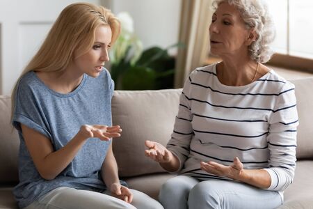 Stressed young blonde grown up daughter arguing with nervous old mature mother, sitting together at home. Irritated elderly woman lecturing adult child, different generations misunderstanding gap.の写真素材