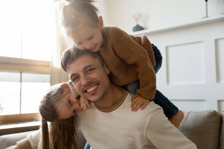 Portrait of excited small preschooler girls kids play with young Caucasian dad relax together in living room, happy father have fun with little daughters enjoy weekend at home, look at camera pictureの写真素材