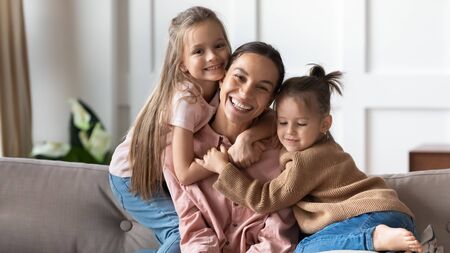 Portrait of overjoyed young mother sit on couch in living room cuddle with little preschooler daughters, happy loving mum pose look at camera relax at home enjoy family weekend with small girl kidsの写真素材