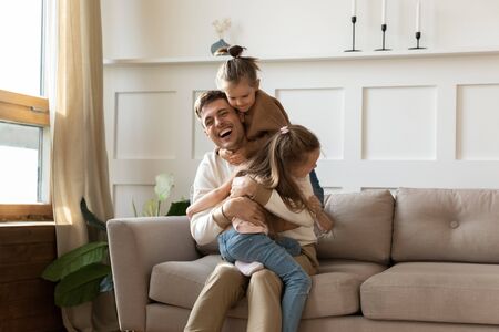 Funny little girls children hug cuddle smiling young Caucasian father sitting relaxing on couch in...