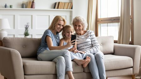 Smiling young attractive woman sitting on comfortable couch with cute little preschool daughter and happy middle aged mother, making selfie photo on mobile phone together in modern living room.の写真素材