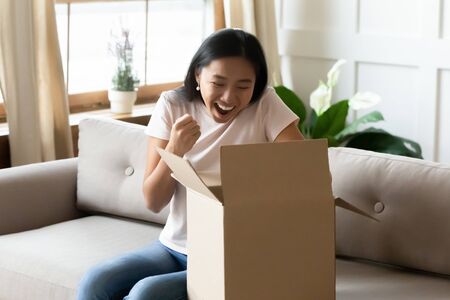 Excited millennial Asian girl sit on couch in living room triumph with online shopping order parcel, overjoyed young Vietnamese woman unpack cardboard box post shipment, good delivery service conceptの写真素材