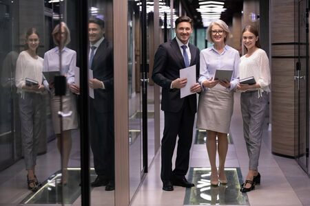 Full length successful smiling different ages business team standing in modern office hall, looking...