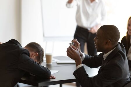 Employee sleeps sit at table put head on hands at morning briefing, african mate takes pictures of...