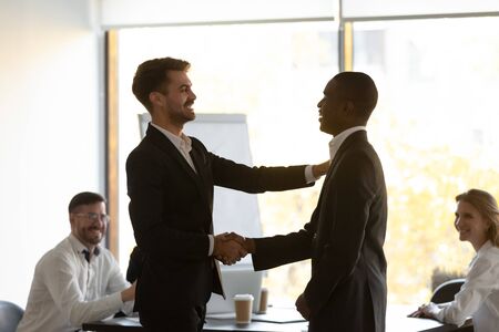 Boss praises and shake hands African member during briefing in office boardroom. Best employee of...