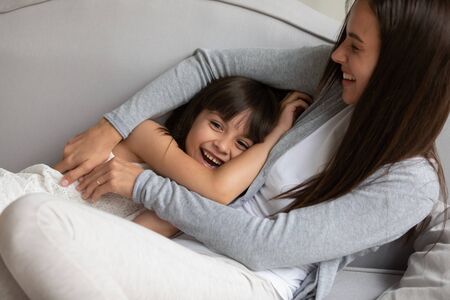 Smiling cute small girl child playing with young mother or nanny cuddle together on couch at home,...