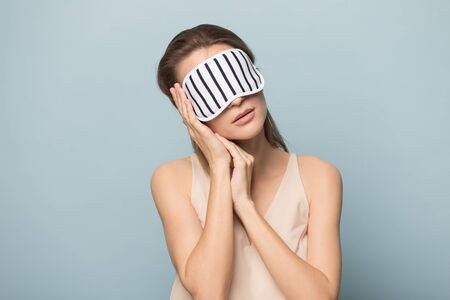 Tired woman wearing sleeping mask isolated on blue studio background relax fall asleep on hands, exhausted calm female in cozy homewear rest sleep standing, dream seeing sweet dreamsの写真素材