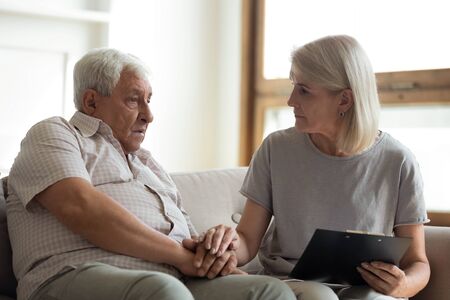 Middle-aged female nurse sit on couch listen to senior male patient complains about health problems, mature woman doctor consult elderly sick man at home show care and support, healthcare conceptの写真素材