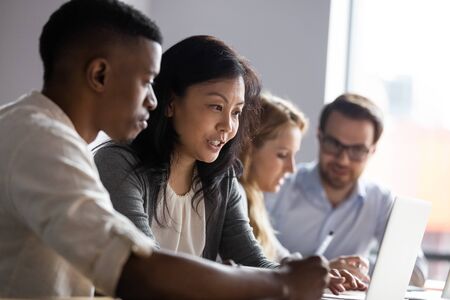 Asian businesswoman mentor teaching African American intern, helping with new corporate software, using laptop, looking at screen, diverse colleagues coworkers working on online project togetherの写真素材