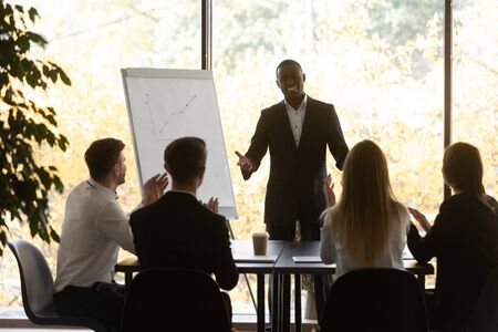 In modern boardroom gathered corporate staff, seated at desk staff members workers applaud expressing appreciation to african male speaker for successful flip chart presentation or educational seminarの写真素材