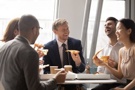 Happy multiethnic employees have fun eating tasty Italian fast food from delivery service, smiling diverse colleagues chat laughing enjoying delicious pizza from takeaway during lunch break in officeの写真素材