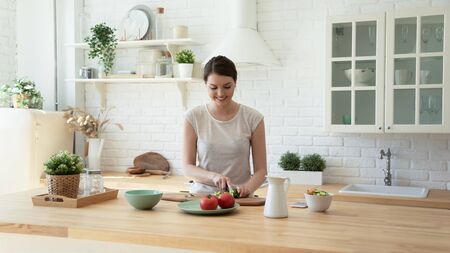 Smiling young woman stand at counter in white modern kitchen cooking healthy breakfast, happy millennial girl housewife preparing food meal, chop vegetables for salad in renovated design homeの写真素材