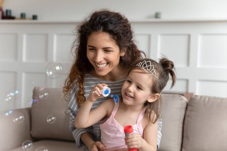 Head shot pleasant young babysitter nanny holding on lap happy small preschool kid girl, blowing soap bubbles indoors. Smiling mommy sitting on couch entertaining with playful little child daughter.の写真素材