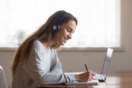 Smiling young woman in Bluetooth earphones make notes in notebook studying on laptop at home, happy...