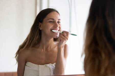 Smiling young woman in towel after shower look in mirror brush clean teeth with toothbrush in bright...