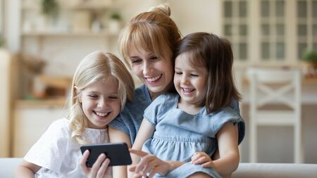 Happy young Caucasian mom and little preschooler daughters relax at home watching video on smartphone together, smiling mother or nanny rest with small little girls children laugh have fun using cellの写真素材