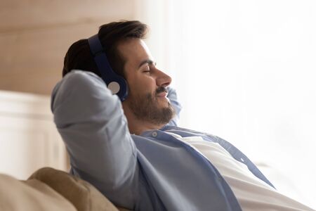 Happy calm millennial Caucasian man relax on comfortable couch with eyes closed listen to music in earphones, smiling young male rest on sofa at home enjoy good quality sound in modern headphonesの写真素材