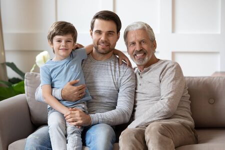 Young smiling handsome father holding on lap small son, sitting together on cozy sofa with middle aged father in living room. Happy three generations male family resting on couch, looking at camera.の写真素材