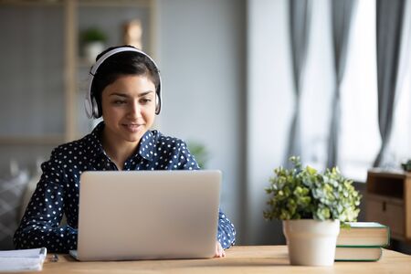 Millennial Indian girl in wireless headphones sit at desk at home working on modern laptop, young...
