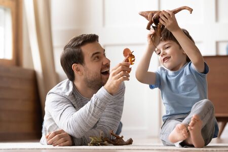 Full length happy young man dad lying on heated warm floor, playing toys with joyful small schoolboy...