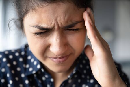 Close up of exhausted unwell millennial Indian girl touch massage temples struggle with migraine or...
