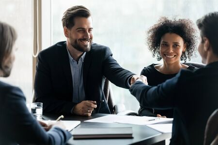 Happy successful businessman in suit shaking hand of business partner at company meeting. Diverse team fasten deal with gesture. Leader in suit with african american assistant congratulates clients.の写真素材