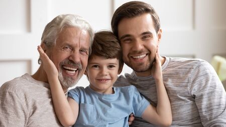 Smiling little schoolboy touching cheeks of cheerful middle aged grandfather and smiling young...