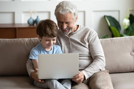 Happy little schoolboy relaxing on couch with smiling hoary senior old grandfather, using computer...