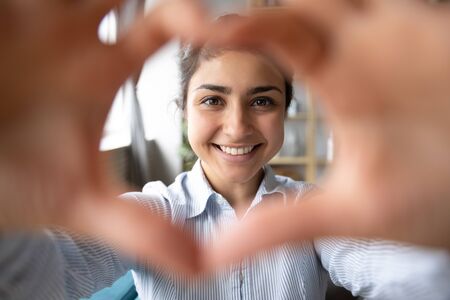Portrait of overjoyed millennial Indian girl have fun make heart with hands posing in living room, profile picture of happy young ethnic woman look at camera smiling relaxing on weekend at homeの写真素材