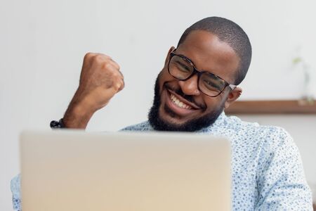 Smiling African American businessman celebrating success, excited by great project results, using laptop, happy employee looking at screen, reading good news, business achievement conceptの写真素材