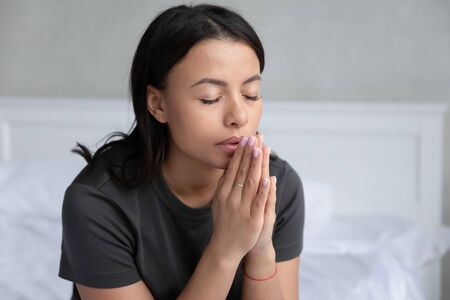 Religious African American young woman sit on bed in bedroom hold hands in prayer ask beg,...