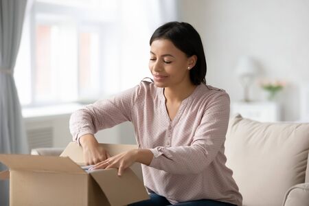 Excited biracial woman sit on couch unpack cardboard box with online order, happy African American...
