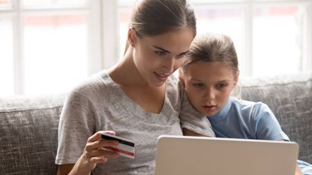 Head shot portrait concentrated young mom with school cute daughter sitting on couch with laptop, making purchases in popular internet store. Smiling woman holding credit card for online payments.の写真素材